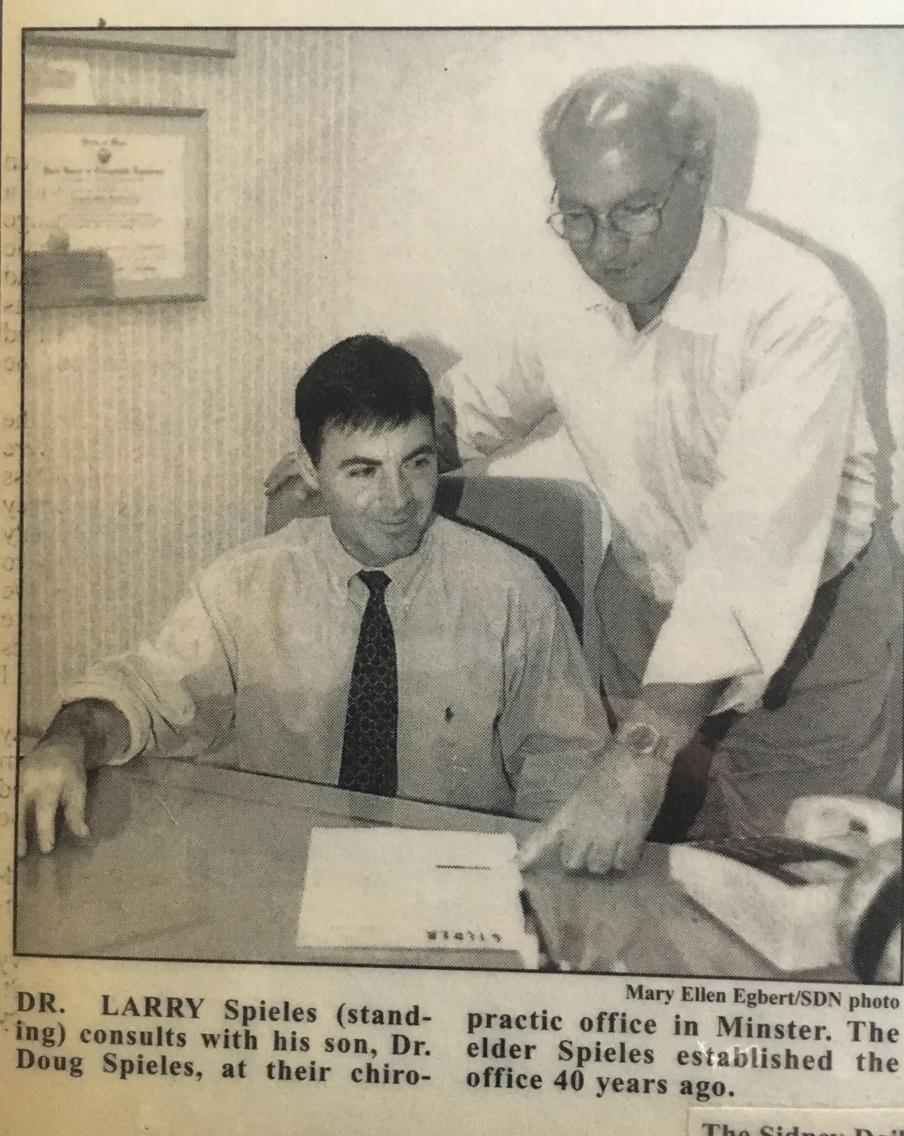Newspaper clipping showing Dr. Larry Spieles standing beside his son Dr. Doug Spieles, seated at a desk.